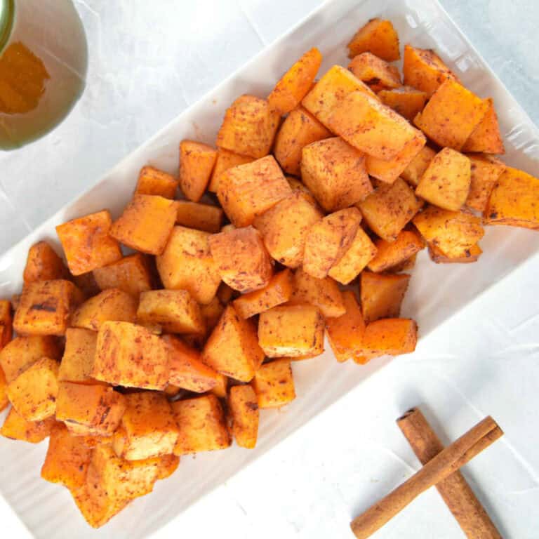 Candied Butternut Squash on a white rectangle plate with two cinnamon sticks to the right. There is a green jar of Maple Syrup a towel with a fall leaves behind it. www.atwistedplate.com