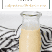 A clear mason jar filled with smooth, golden Paleo mustard sauce on a wooden board next to fresh herbs.