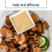 Golden-brown crispy marinated pork cubes on a white plate, garnished with fresh lime wedges and a small bowl of yellow mustard dipping sauce..