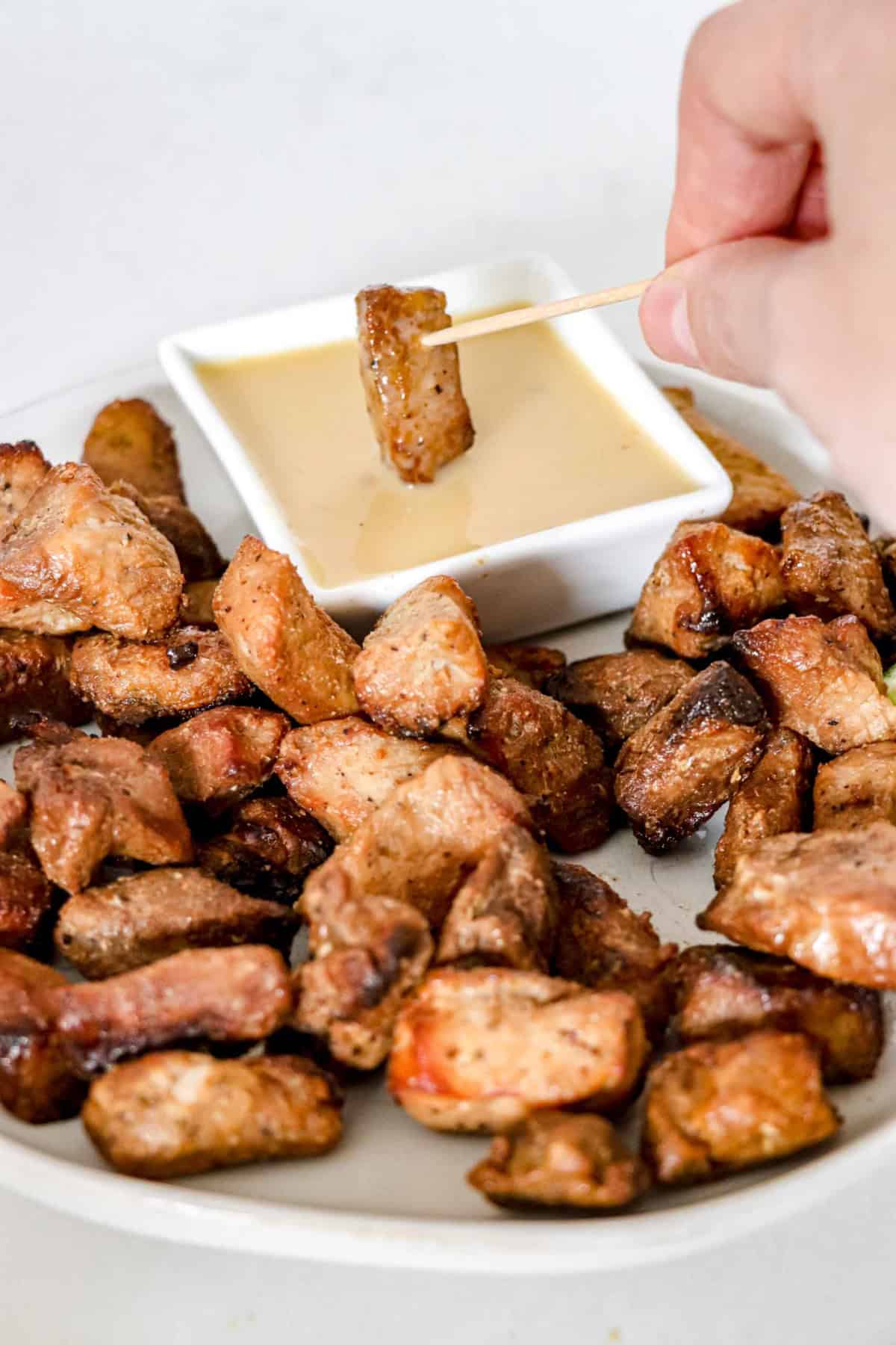 Side angle of Pork Bites on a white round plate with cut up limes and a square bowl of Paleo Mustard Sauce with a pork bite being dipped in. www.atwistedplate.com/pork-bites/