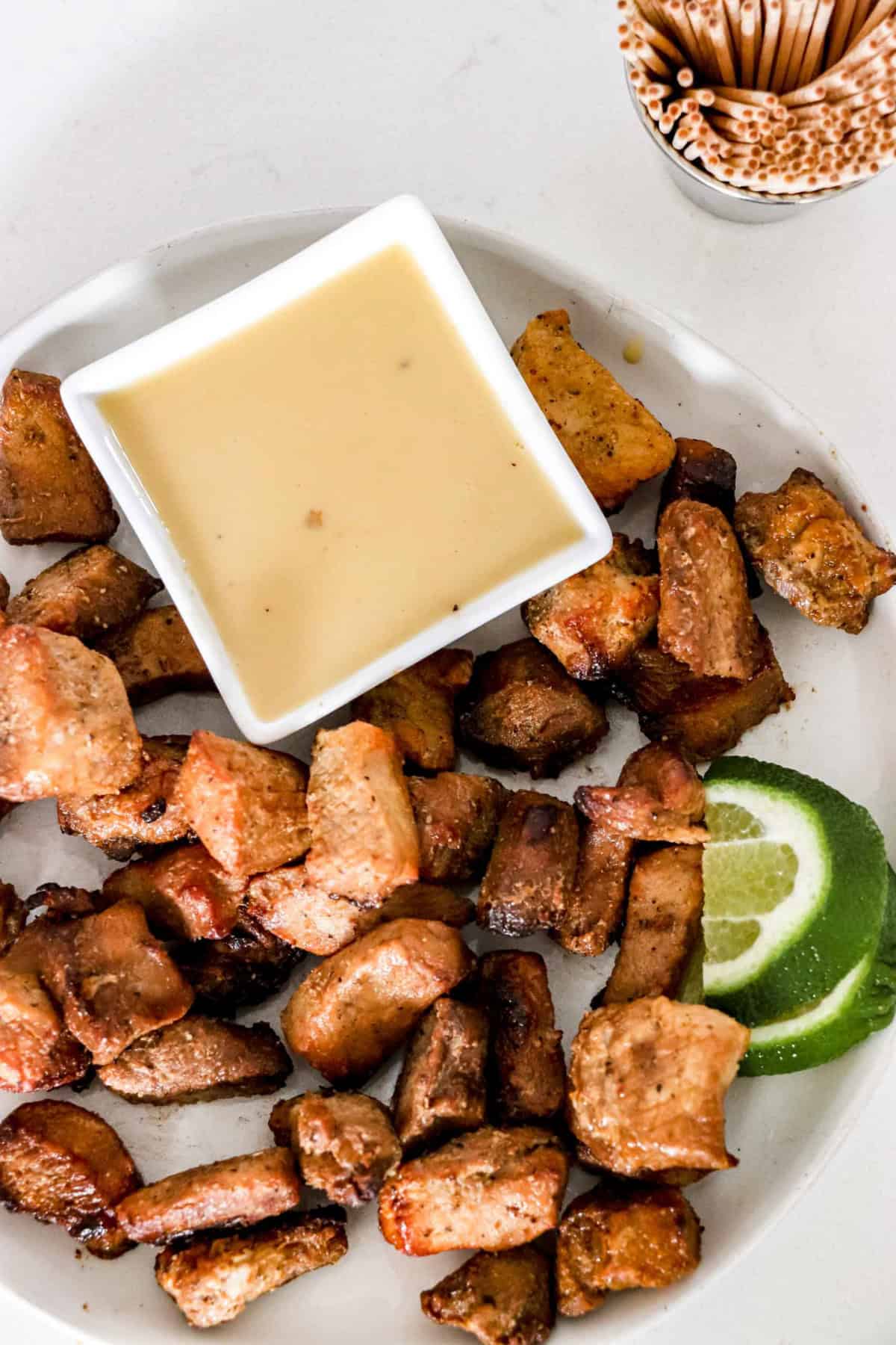 Pork Bites on a white round plate with cut up limes and a square bowl of Paleo Mustard Sauce. On the sider there is a container of toothpicks. www.atwistedplate.com/pork-bites/