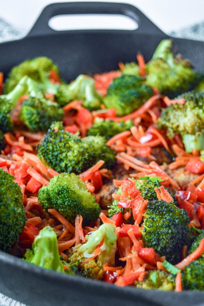 Angled, close up side view of a cast iron skillet with chicken, broccoli, carrots, and red peppers.  Cast iron is sitting on a teal and white towel. www.atwistedplate.com