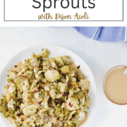 Shaved and roasted crispy Brussel sprouts on a white plate, drizzled with a creamy yellow Dijon aioli sauce and sprinkled with red pepper flakes