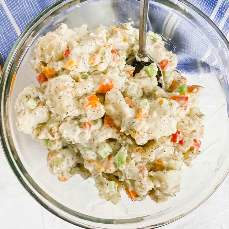 Top view of roasted pepper tomato salad In a glass bowl with peppers below the bowl. There is a blue and white towel at the top. www.atwistedplate.com