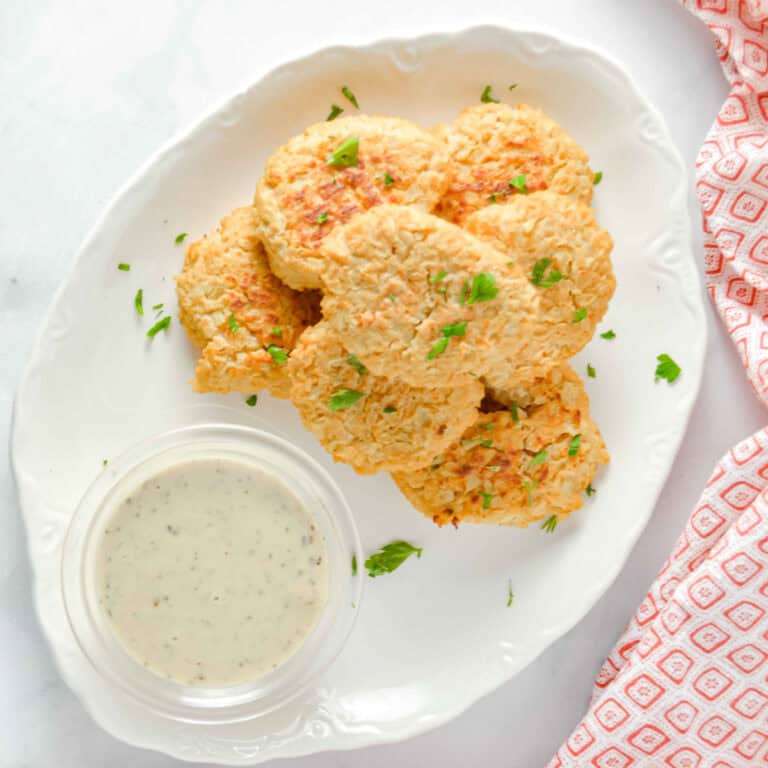 Top view Cheesy Cauliflower Bites on a white plate with a cup of ranch dressing. There is a red and white towel in the background. www.atwistedplate.com/cheesy-cauliflower-bites/