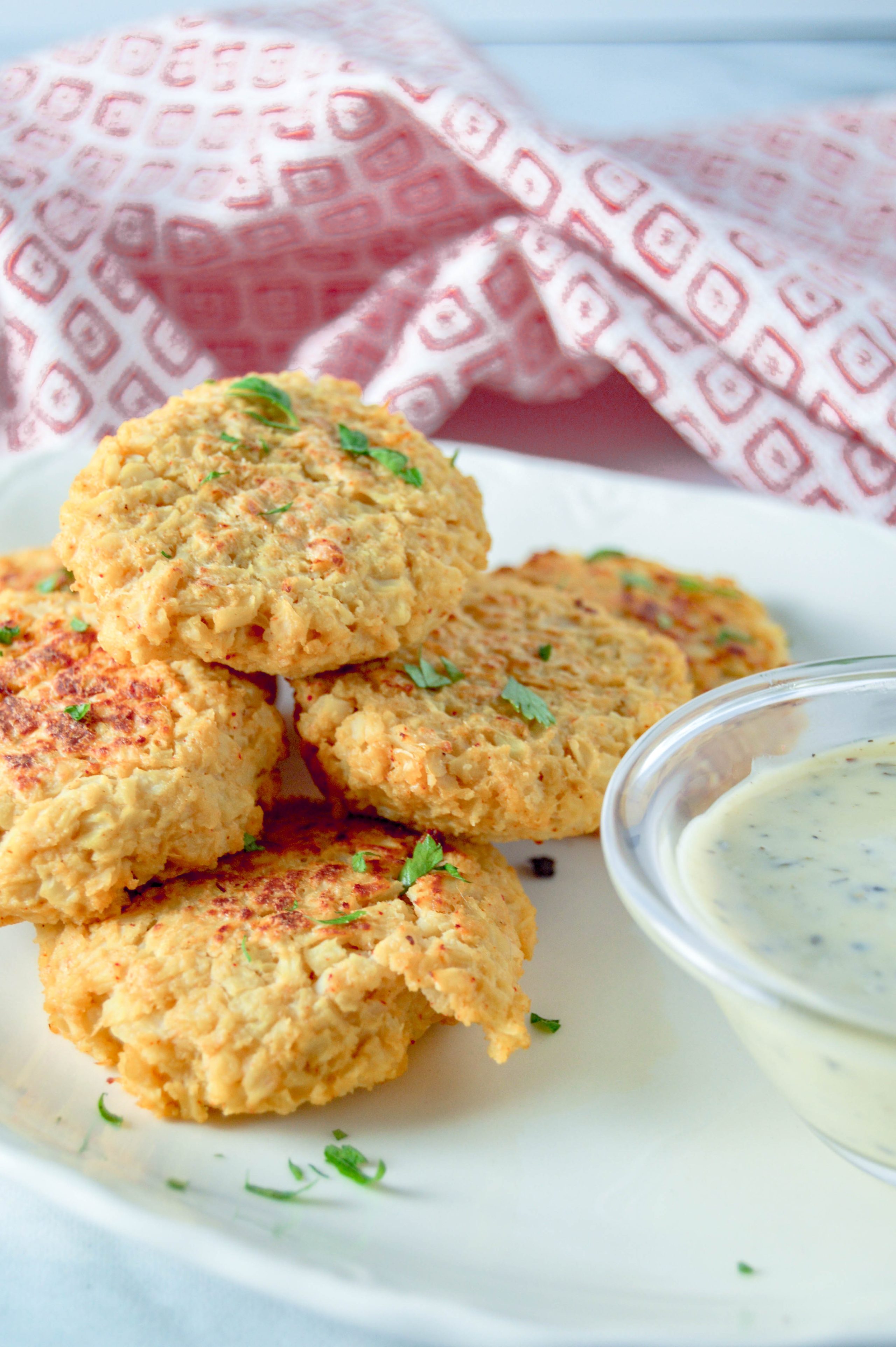 Cheesy Vegan Cauliflower Bites on a white plate with a cup of ranch dressing. There is a red and white towel in the background. www.atwistedplate.com/cheesy-cauliflower-bites/