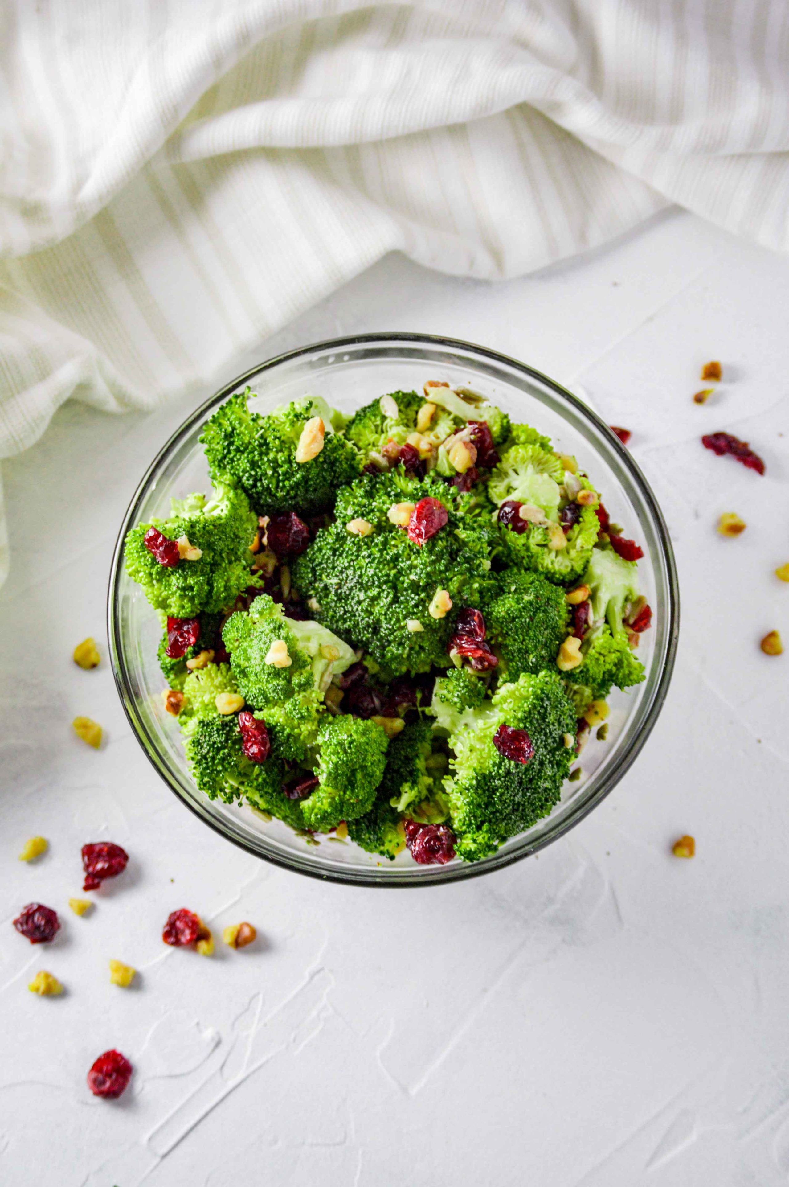 Top view of Broccoli salad in a clear bowl against a white background. there are walnuts and dried cranberries scattered around and a white towel at the top of the image. https://www.atwistedplate.com/broccoli-salad/