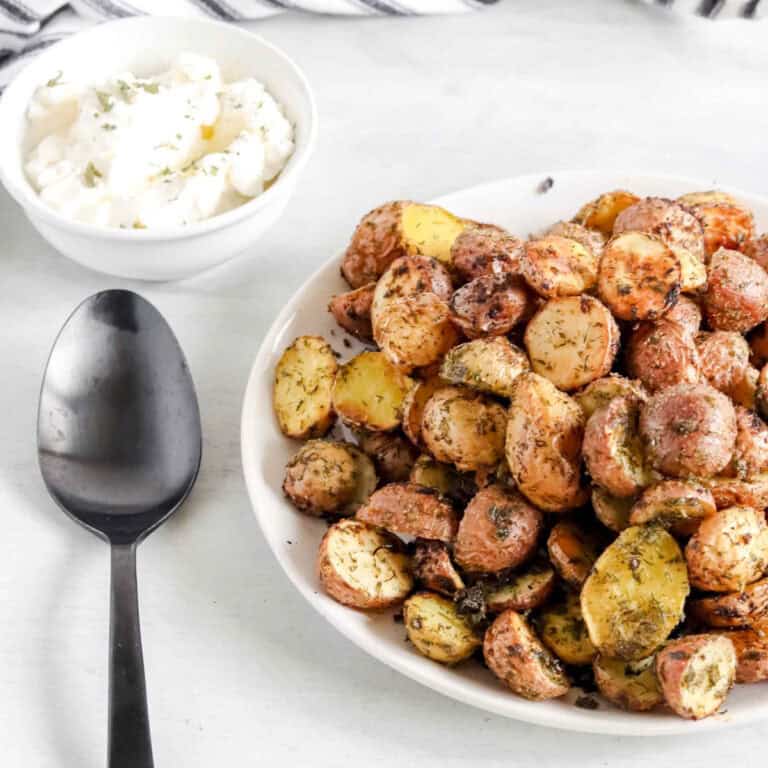 Top view of Roasted Ranch Potatoes on a white plate with a bowl of sour cream behind. https://www.atwistedplate.com/roasted-ranch-potatoes/