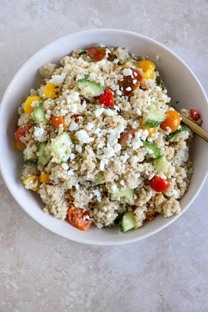 A vibrant bowl of fluffy quinoa mixed with diced red tomatoes, crisp green cucumbers, and crumbled white feta cheese, drizzled with herb-flecked Italian dressing