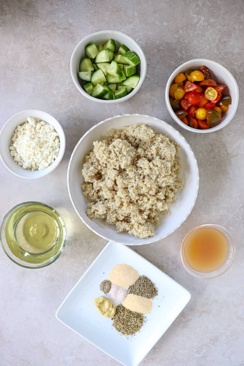 Ingredients including a bowl of fluffy quinoa mixed with diced red tomatoes, crisp green cucumbers, and crumbled white feta cheese, drizzled with herb-flecked Italian dressing