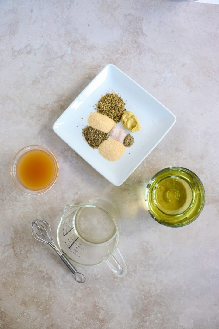 Ingredients for Italian dressing including herbs, apple cider vinegar, olive oil measuring cup and whisk.