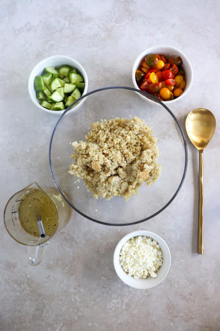 Ingredients including a bowl of fluffy quinoa mixed with diced red tomatoes, crisp green cucumbers, and crumbled white feta cheese, drizzled with herb-flecked Italian dressing