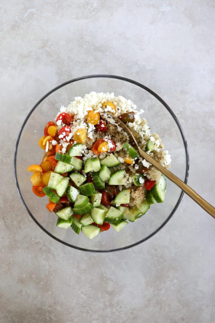 A vibrant bowl of fluffy quinoa mixed with diced red tomatoes, crisp green cucumbers, and crumbled white feta cheese, drizzled with herb-flecked Italian dressing