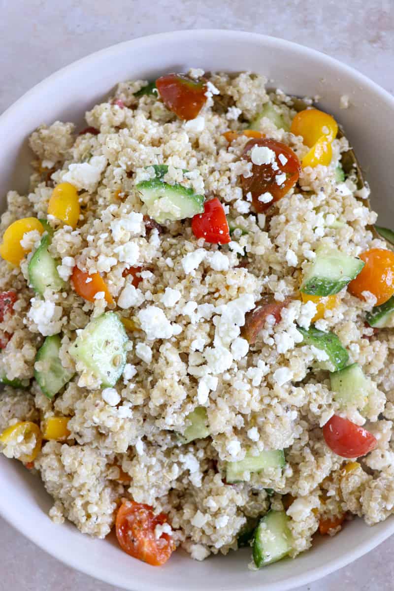 A vibrant bowl of fluffy quinoa mixed with diced red tomatoes, crisp green cucumbers, and crumbled white feta cheese, drizzled with herb-flecked Italian dressing