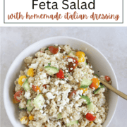 A vibrant bowl of fluffy quinoa mixed with diced red tomatoes, crisp green cucumbers, and crumbled white feta cheese, drizzled with herb-flecked Italian dressing