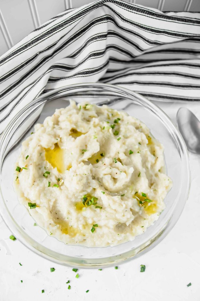 A top view of Garlic and Chive Mashed Cauliflower in a clear bowl with melted ghee and chive on top against a white background. There is a black and white towel in the background and silver spoon to the left. https://www.atwistedplate.com/garlic-&-chive-mashed-cauliflower/