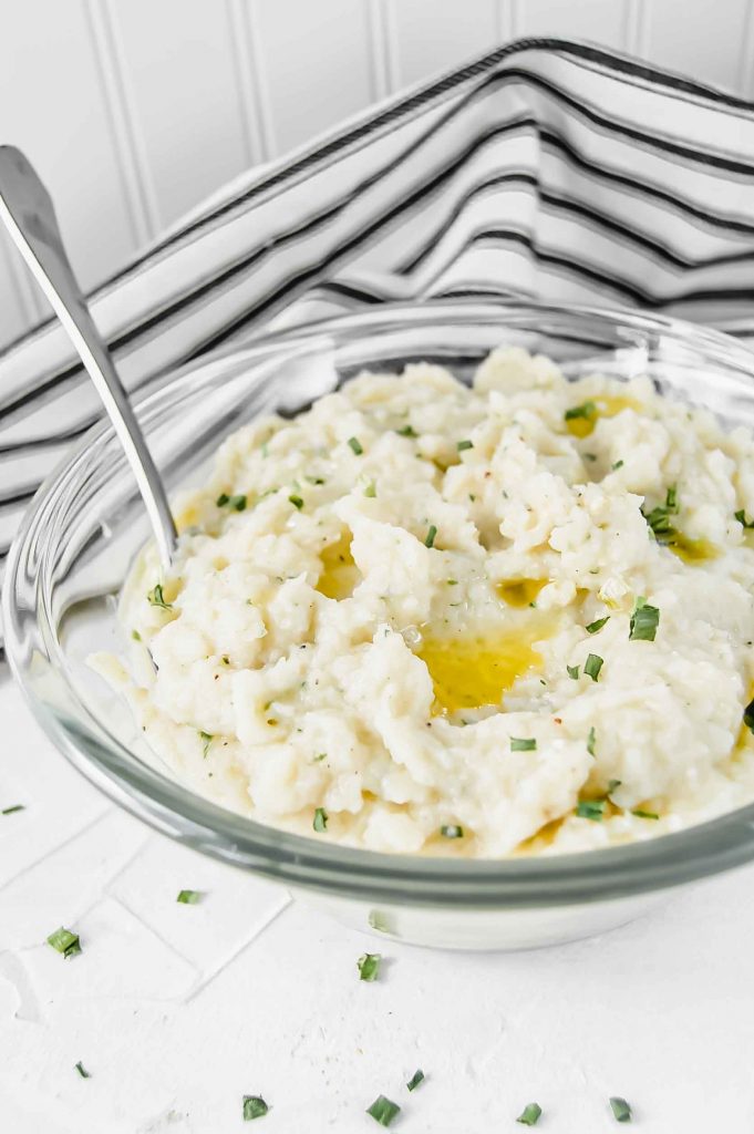 An angled view of Garlic and Chive Mashed Cauliflower in a clear bowl with melted ghee and chive on top against a white background. There is a black and white towel in the background and silver spoon to the left. https://www.atwistedplate.com/garlic-&-chive-mashed-cauliflower/