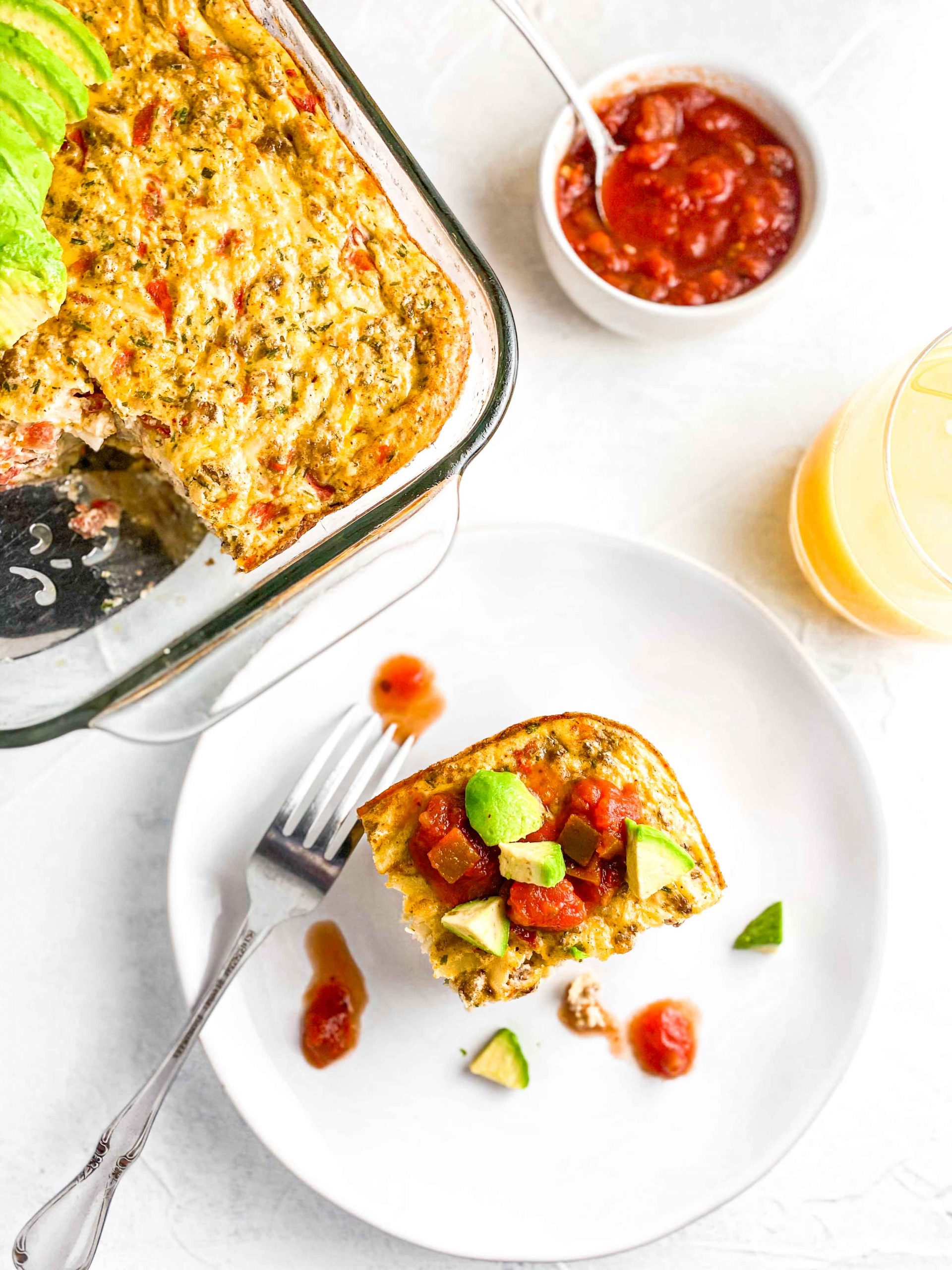 Easy Potato and Sausage Bake on a white plate topped with Avocado and Salsa with the casserole, bowl of salsa and Orange Juice against a white background.  https://www.atwistedplate.com/easy-potato-and-sausage-bake/