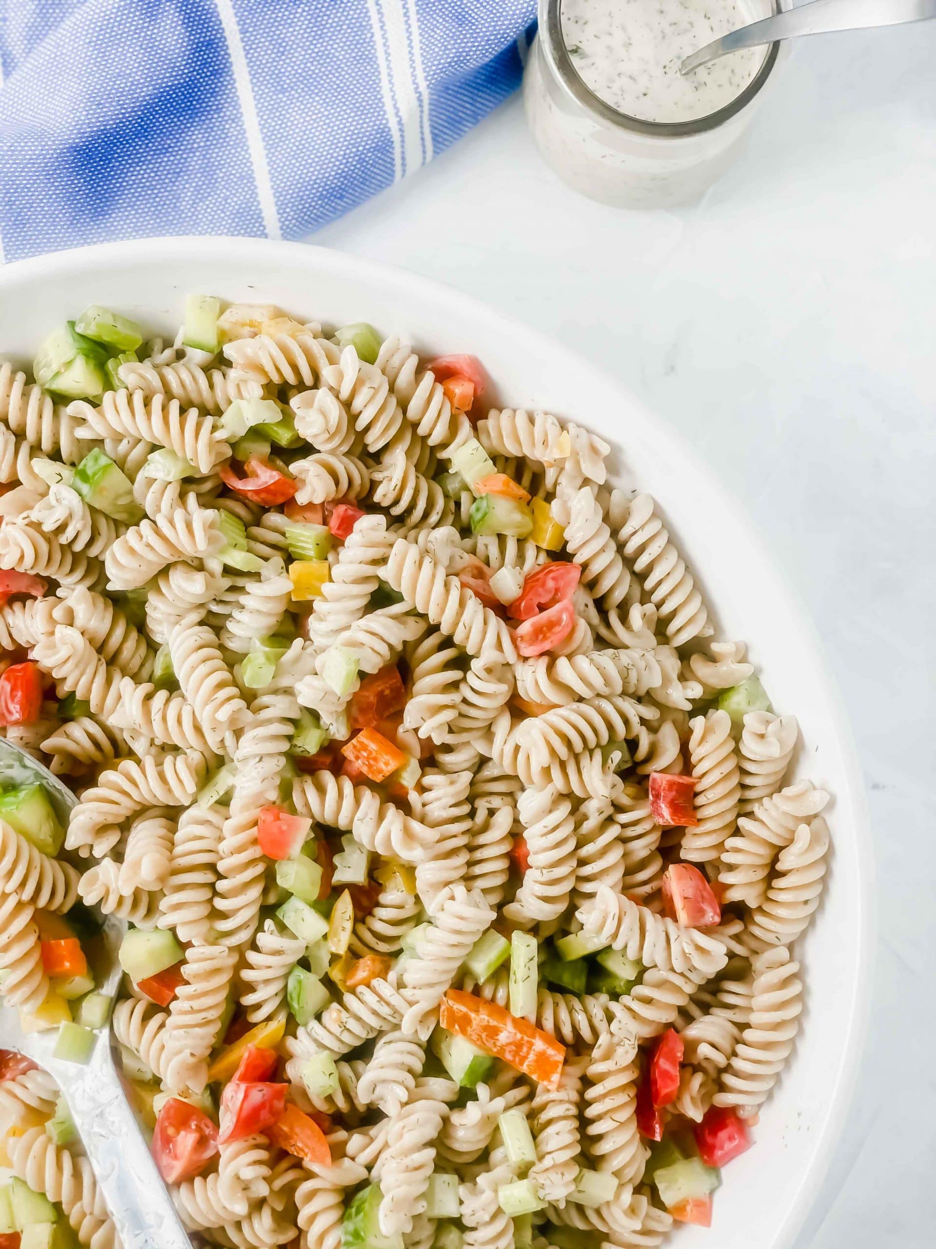 Image of Dill pasta salad in a white bowl. There is a jar of creamy dill pasta sauce and a spoon. At the top there blue bowl with a white towel. https://www.atwistedplate.com/dill-pasta-salad/