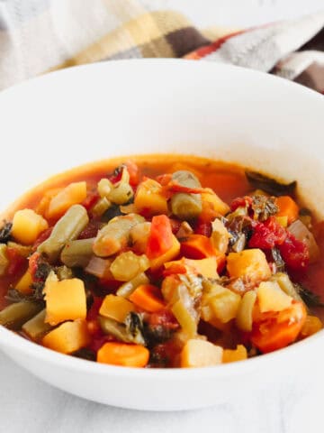 A Bowl of Harvest Veggie Soup against a Marble background with two spoons stacked inbetween. Behind the bowls is fall colored plaid towel. https://www.atwistedplate.com/harvest-veggie-soup/