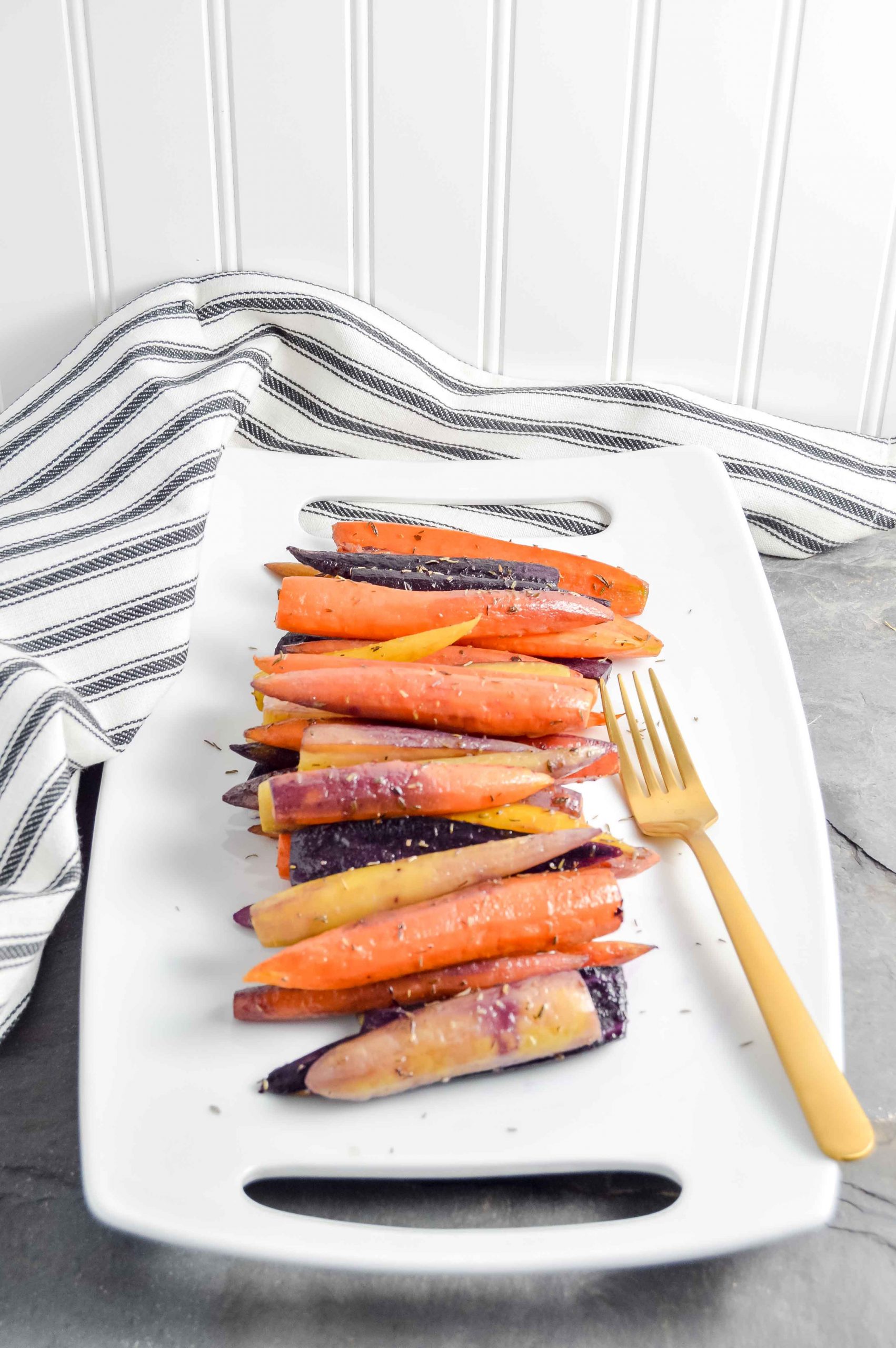 Image of a side angle view of Perfectly Roasted Carrots on a rectangle white plate with a gold fork.  The plate sites on a gray blue background with a black and white striped towel in the top left corner.   https://www.atwistedplate.com/perfectly-roasted-carrots/