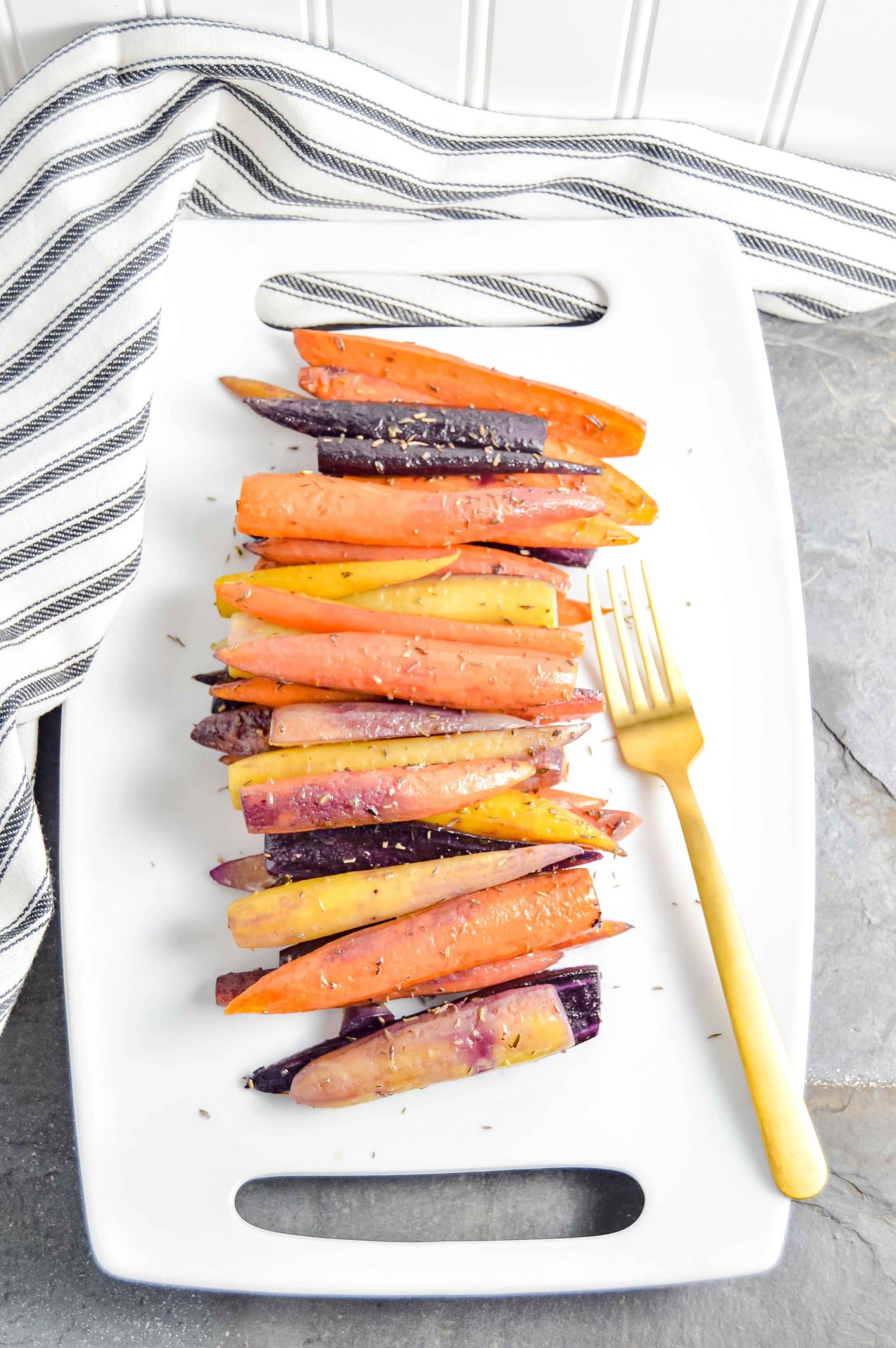 Image of a side angle view of Perfectly Roasted Carrots on a rectangle white plate with a gold fork. The plate sites on a gray blue background with a black and white striped towel in the top left corner. https://www.atwistedplate.com/perfectly-roasted-carrots/