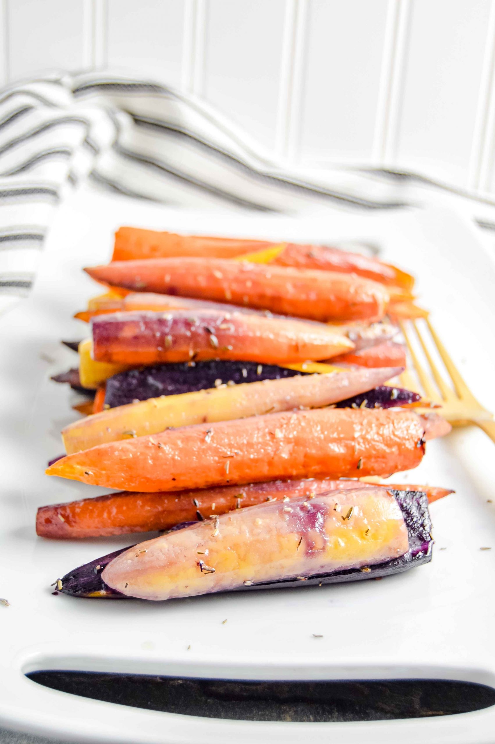 Image of a side angle view of Perfectly Roasted Carrots on a rectangle white plate with a gold fork.  The plate sites on a gray blue background with a black and white striped towel in the top left corner.   https://www.atwistedplate.com/perfectly-roasted-carrots/