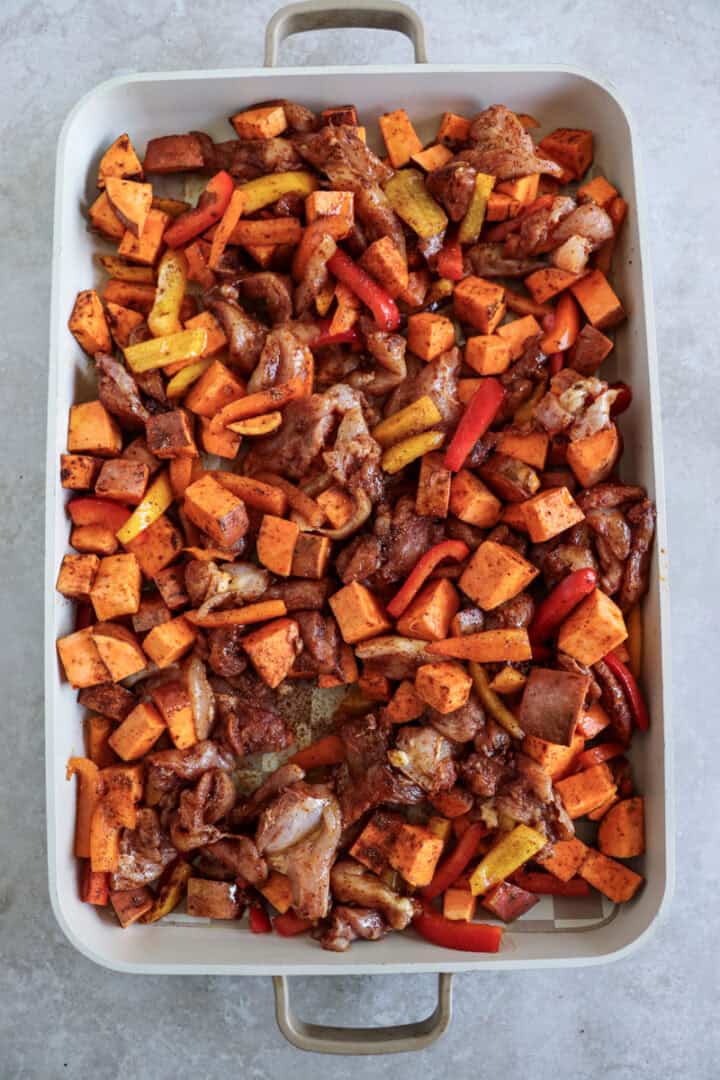 A large sheet pan filled with roasted chicken thigh strips, sliced bell peppers, red onions, and cubed sweet potatoes, all seasoned with smoky fajita spices.