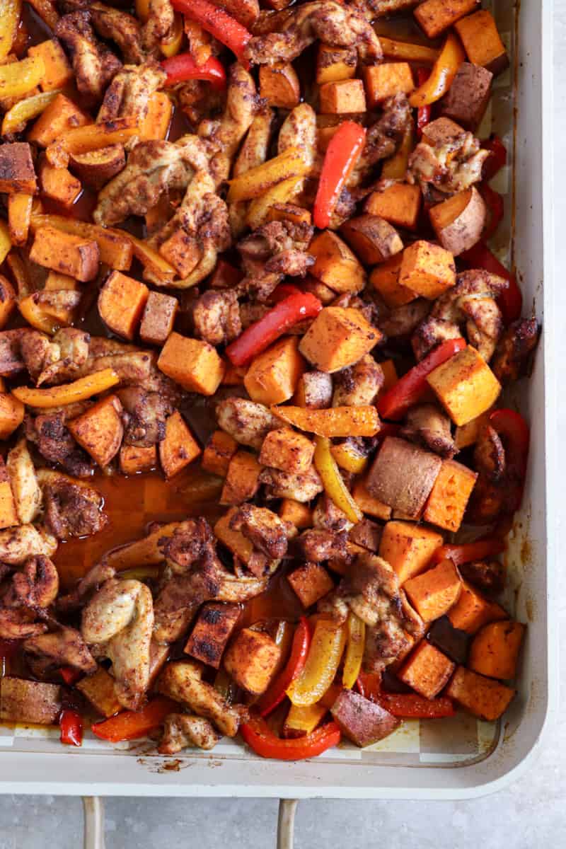 A large sheet pan filled with roasted chicken thigh strips, sliced bell peppers, red onions, and cubed sweet potatoes, all seasoned with smoky fajita spices.
