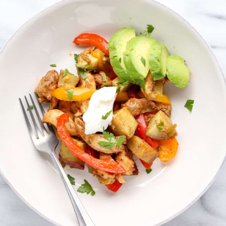 A large sheet pan filled with roasted chicken thigh strips, sliced bell peppers, red onions, and cubed sweet potatoes, all seasoned with smoky fajita spices.