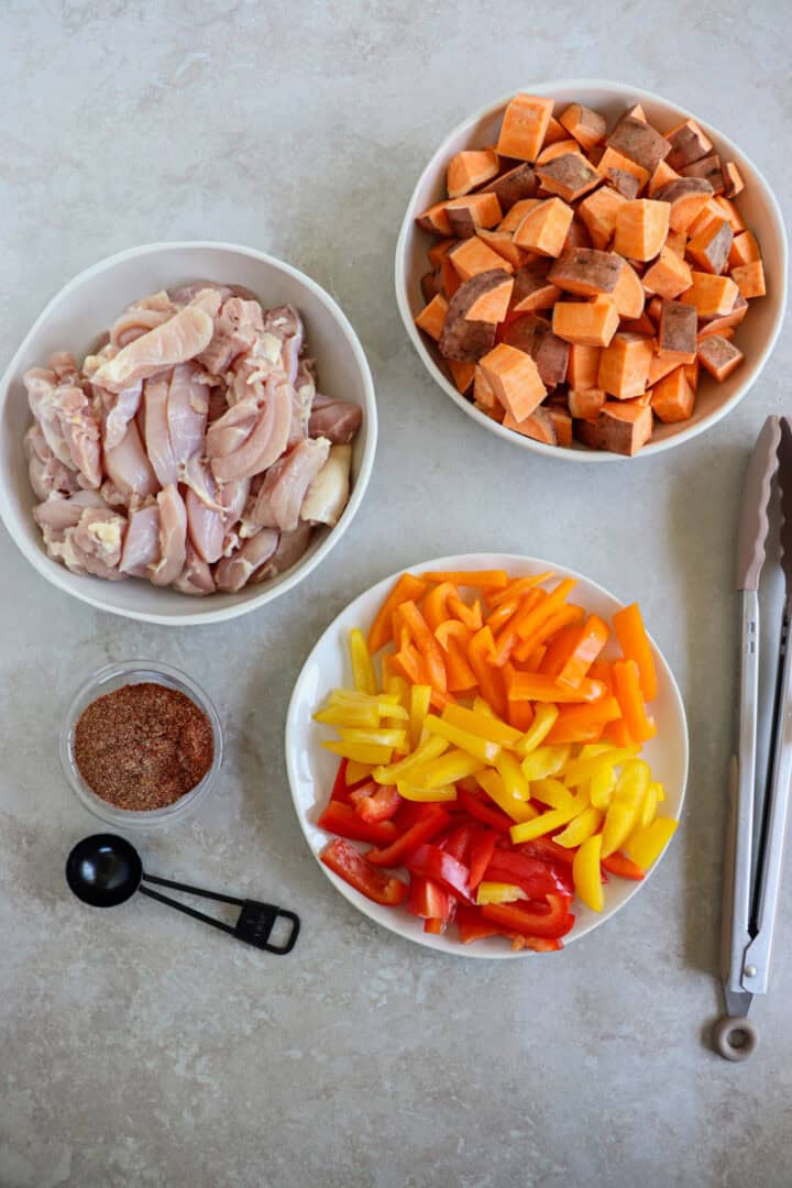 A large sheet pan filled with roasted chicken thigh strips, sliced bell peppers, red onions, and cubed sweet potatoes, all seasoned with smoky fajita spices.