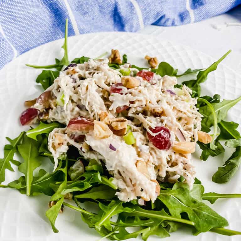 Side view of Chicken Walnut and Grape Salad on a bed of Arugula on a white plate. There is a blue and white towel along the top with Grapes and Walnuts along the bottom of the image. https://www.atwistedplate.com/chicken-walnut-and-grape-salad/
