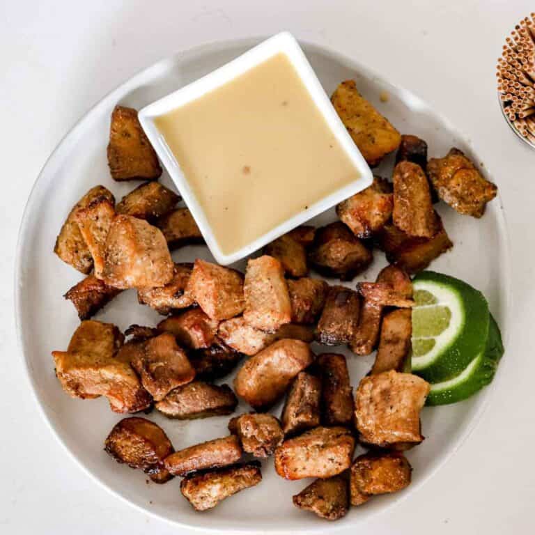 Pork Bites on a white round plate with cut up limes and a bowl of Paleo Mustard Sauce. www.atwistedplate.com/pork-bites/
